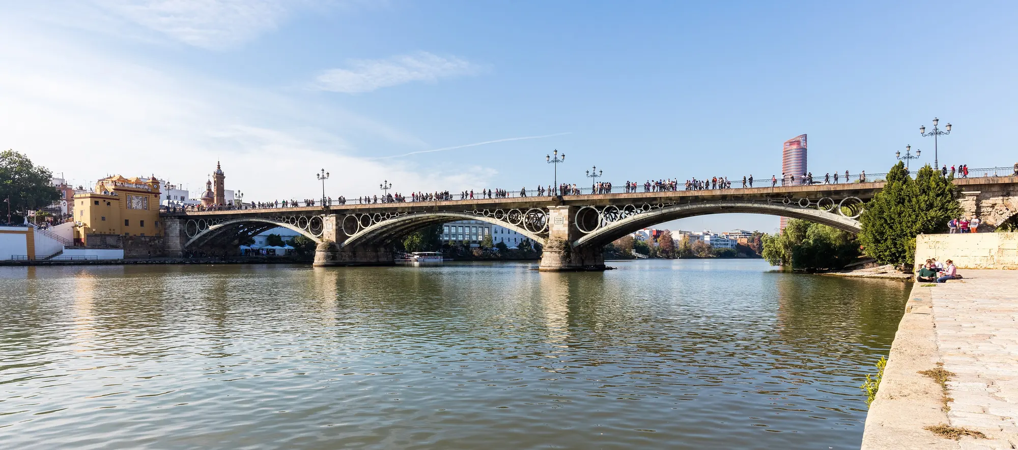Železný most Isabel II. překlenující Guadalquivir v Seville, s Trianskou žlutou kaplí a věžemi na levém břehu a lidmi přecházejícími přes most