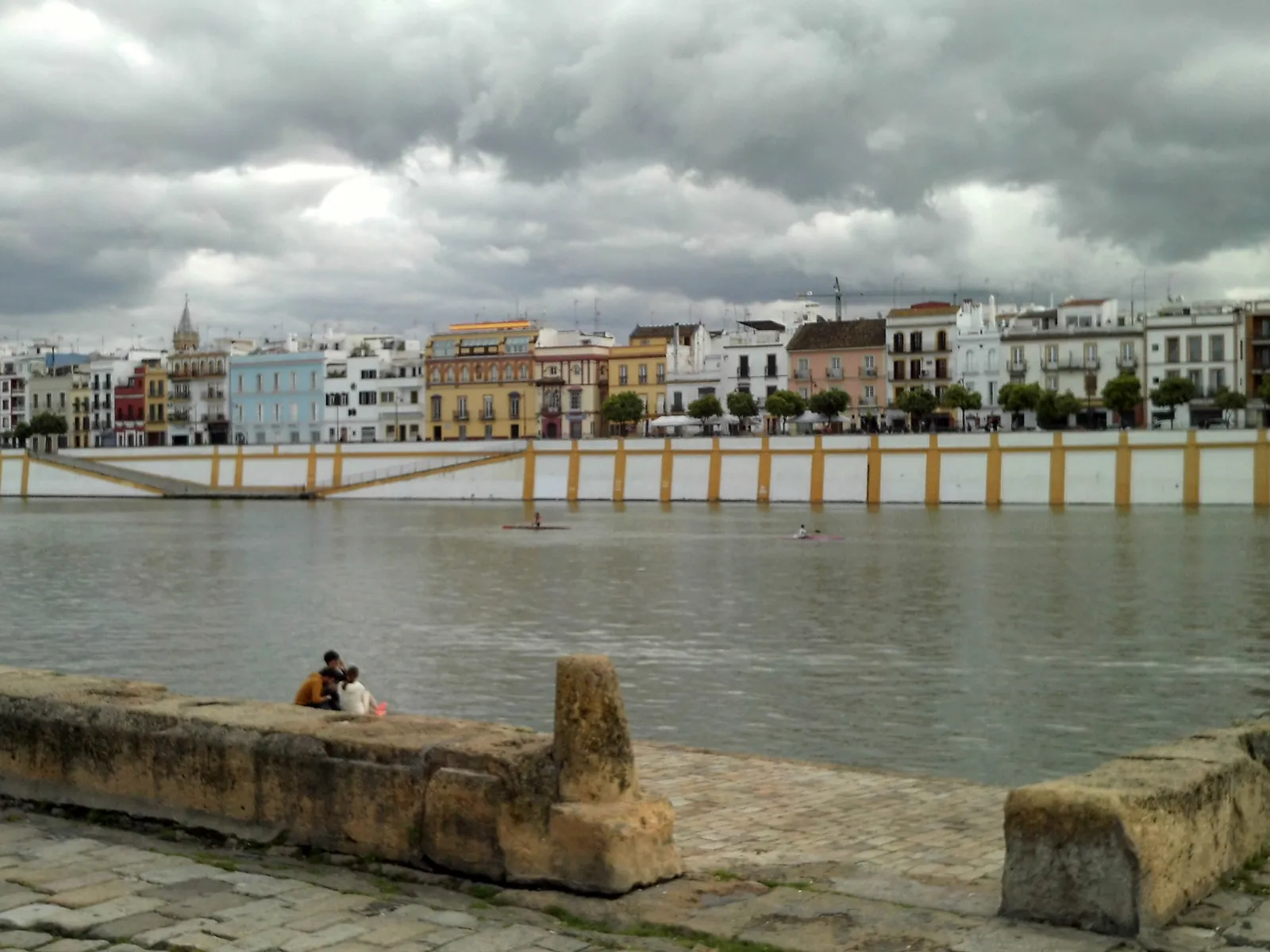 Bílé a barevné domy na Calle Betis podél řeky, pohled z protějšího břehu pod zataženou oblohou