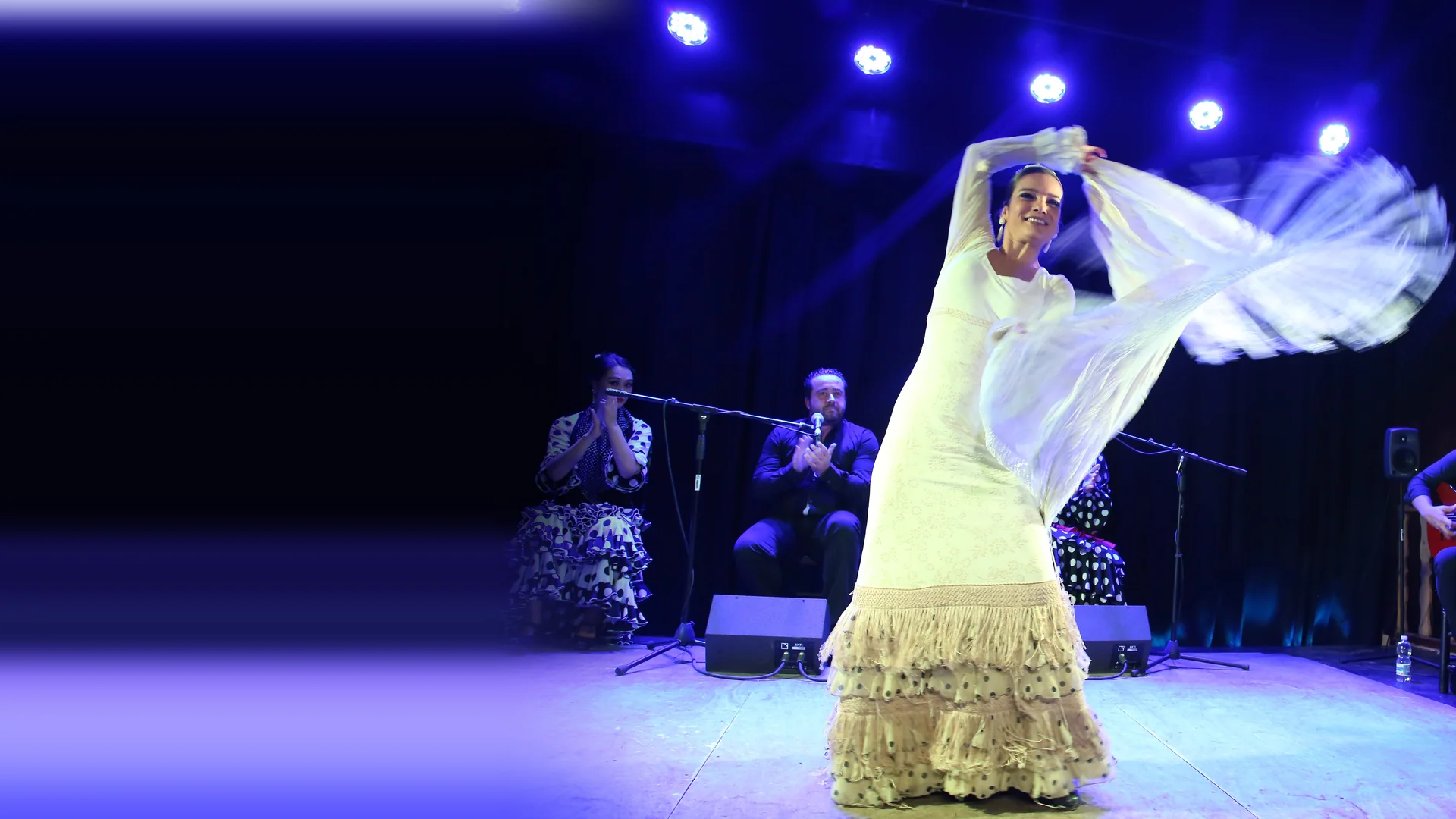 Flamenková tanečnice v bílých šatech rozvíří svůj šál na jevišti zalitém modrým světlem, za ní sedí zpěvák, umělec doprovázející tleskáním a kytarista